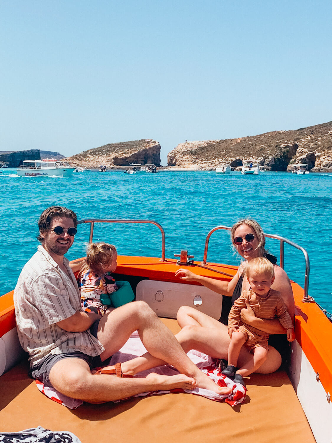 Man, woman and two children sit on small orange boat in bright blue water with rocky island in the distance