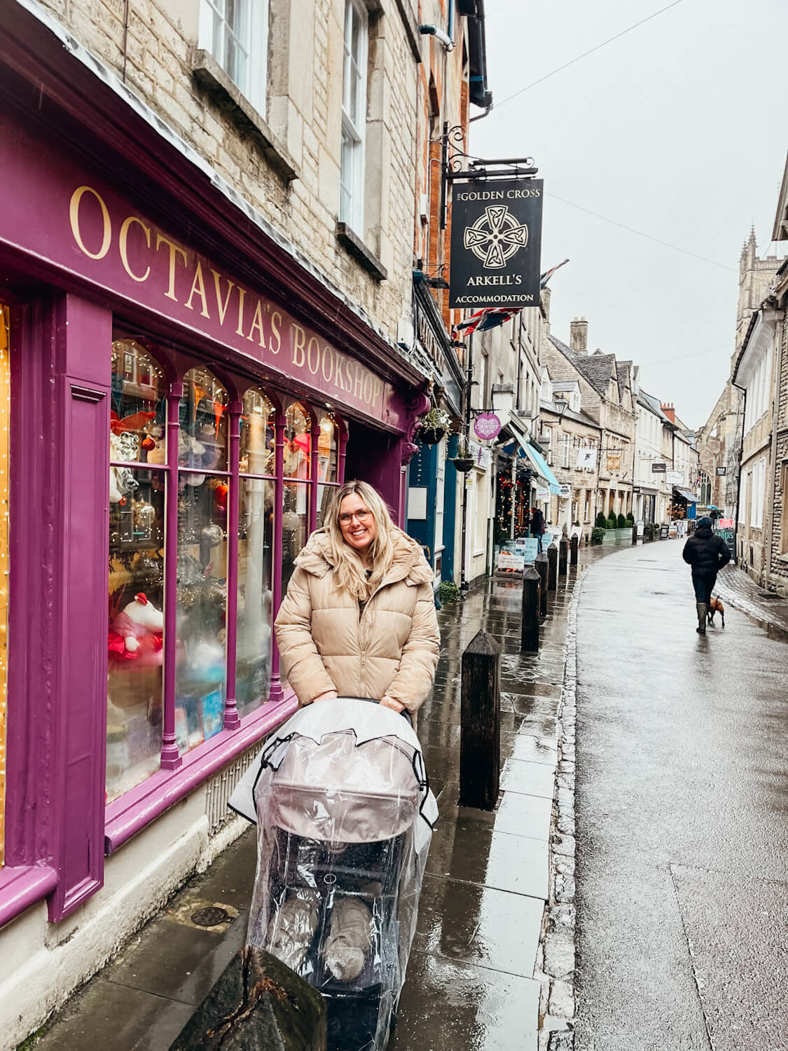 Woman stands with stroller in front of bookshop with a wooden purple front on an old shopping road in Cirencester, Cotswolds