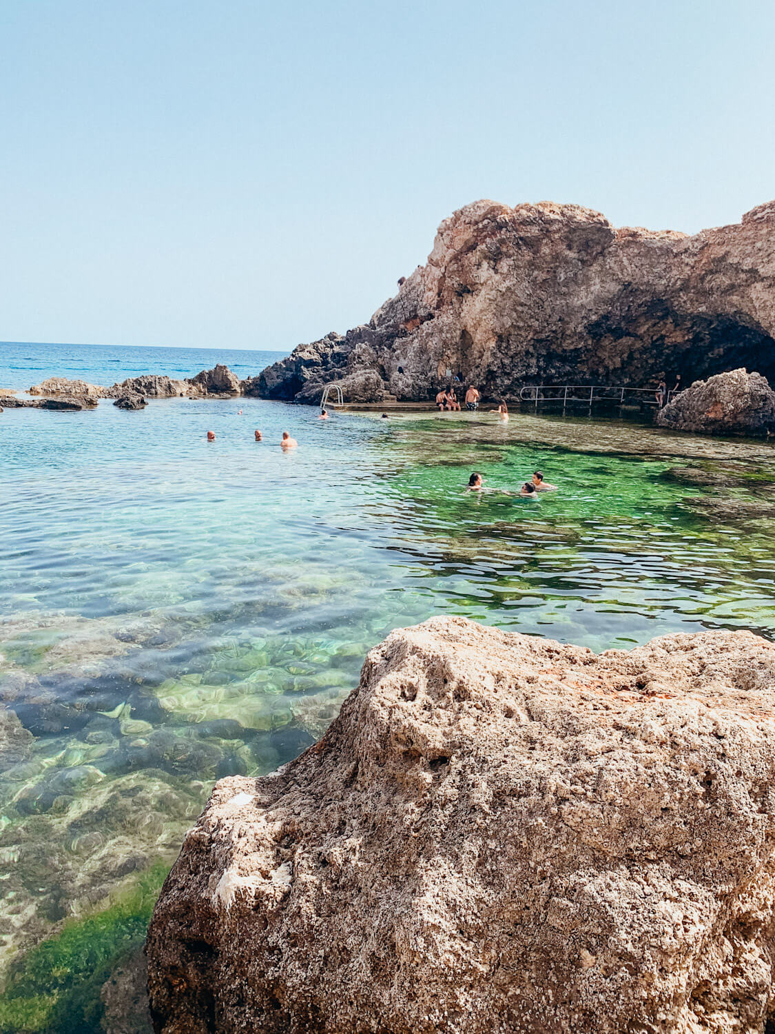People float in clear green water surrounded by rocks at Ghar Lapsi in Malta