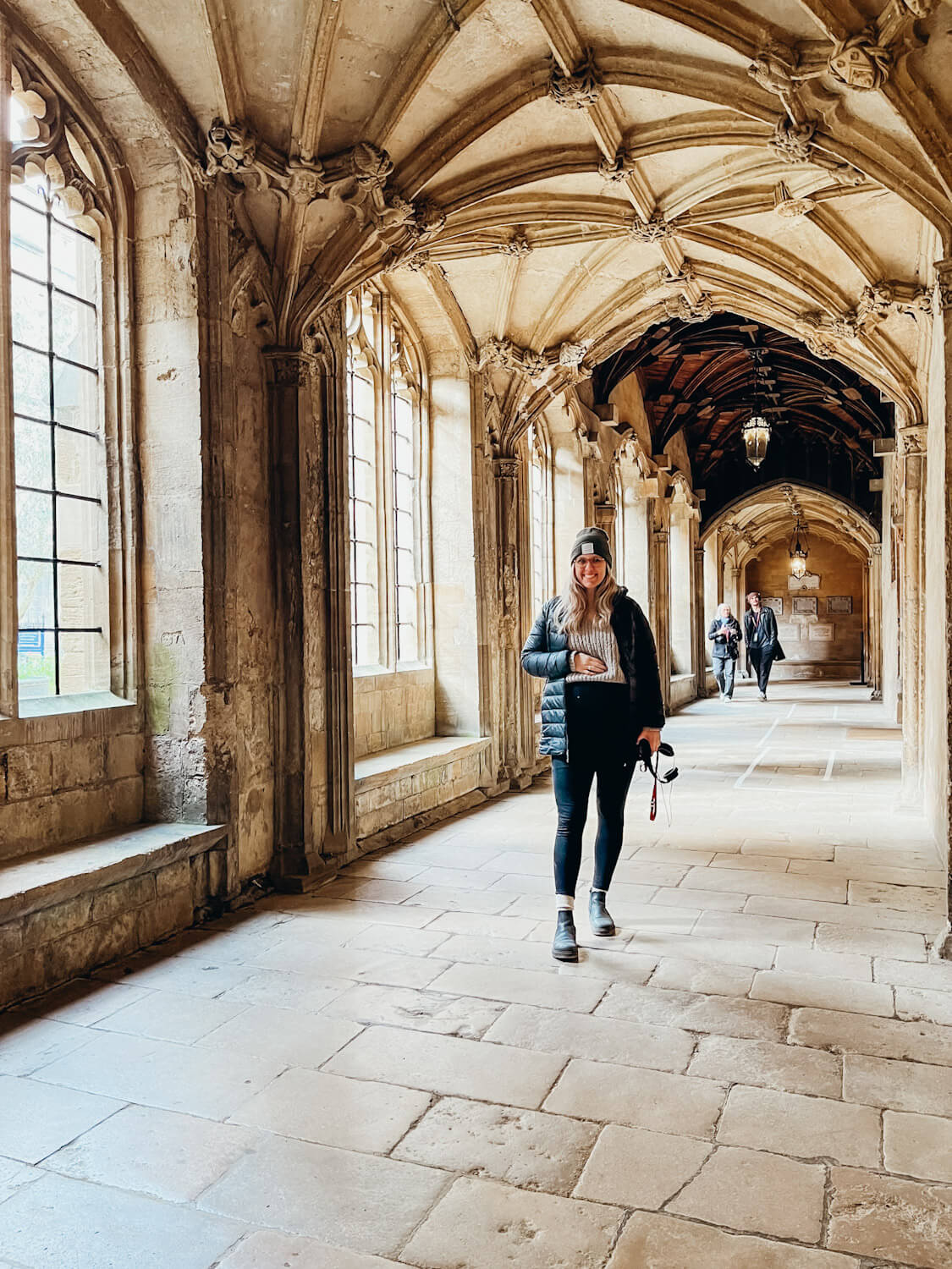 Woman stands on stone pathway under stone arches next to large windows at Christ Church College in Oxford