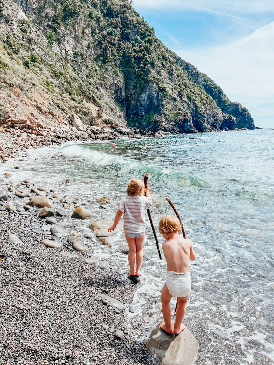 Two kids walk with sticks on rocky beach in Riomaggiore Cinque Terre Italy