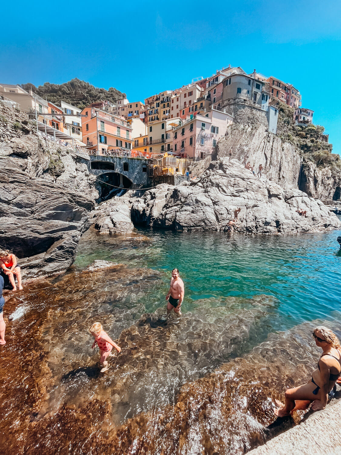 People walk out of harbor water with colorful buildings on the hill above them in the Cinque Terre