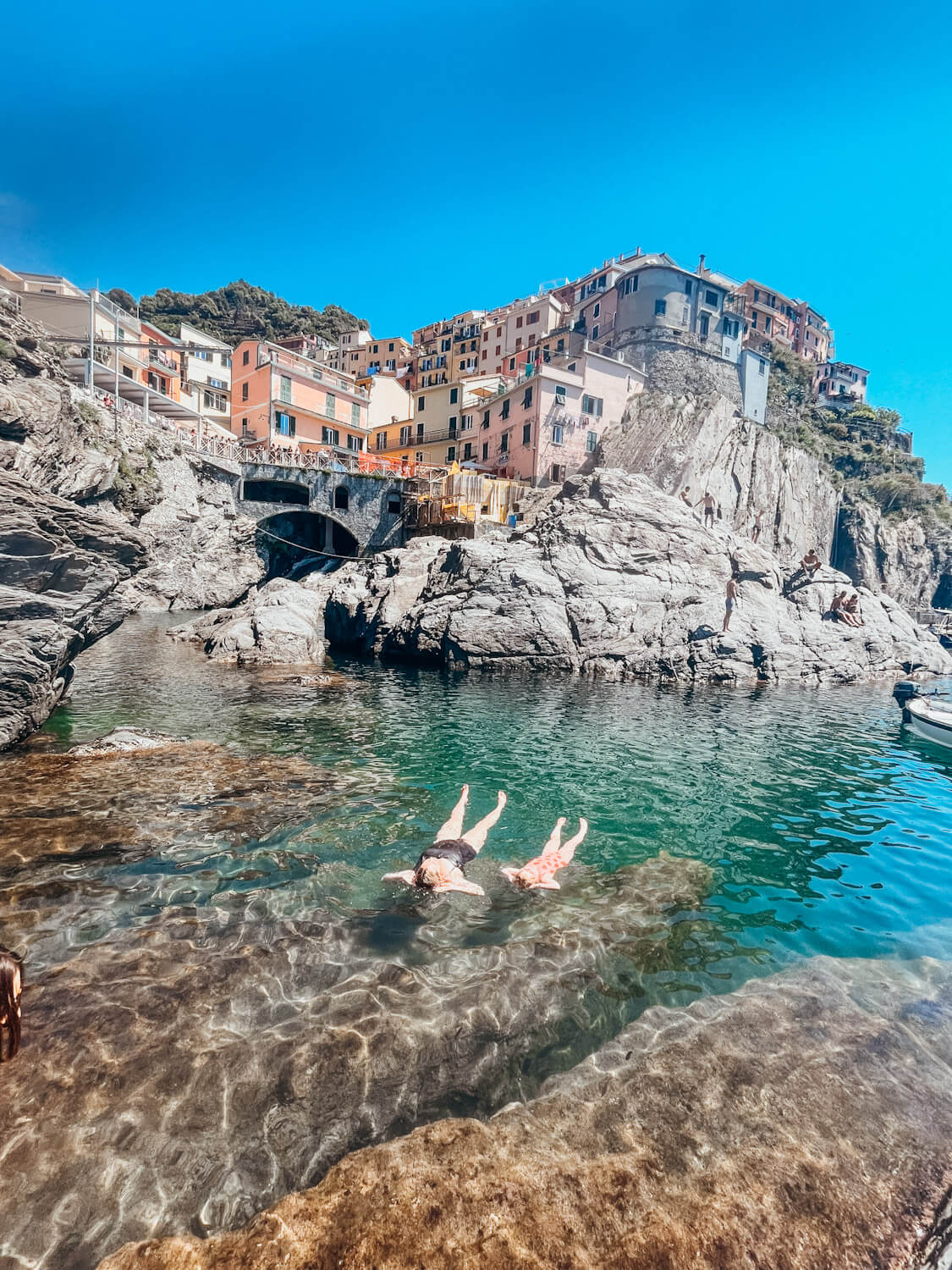 Two people float on their backs in harbor with colorful buildings above them on the hill