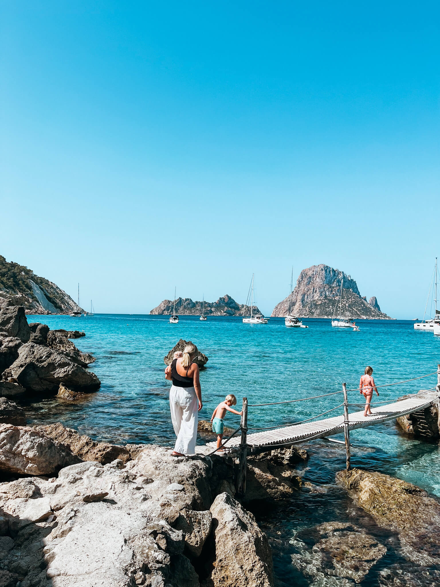 Woman and chidren walk across wooden dock over clear blue water in Ibiza as a family