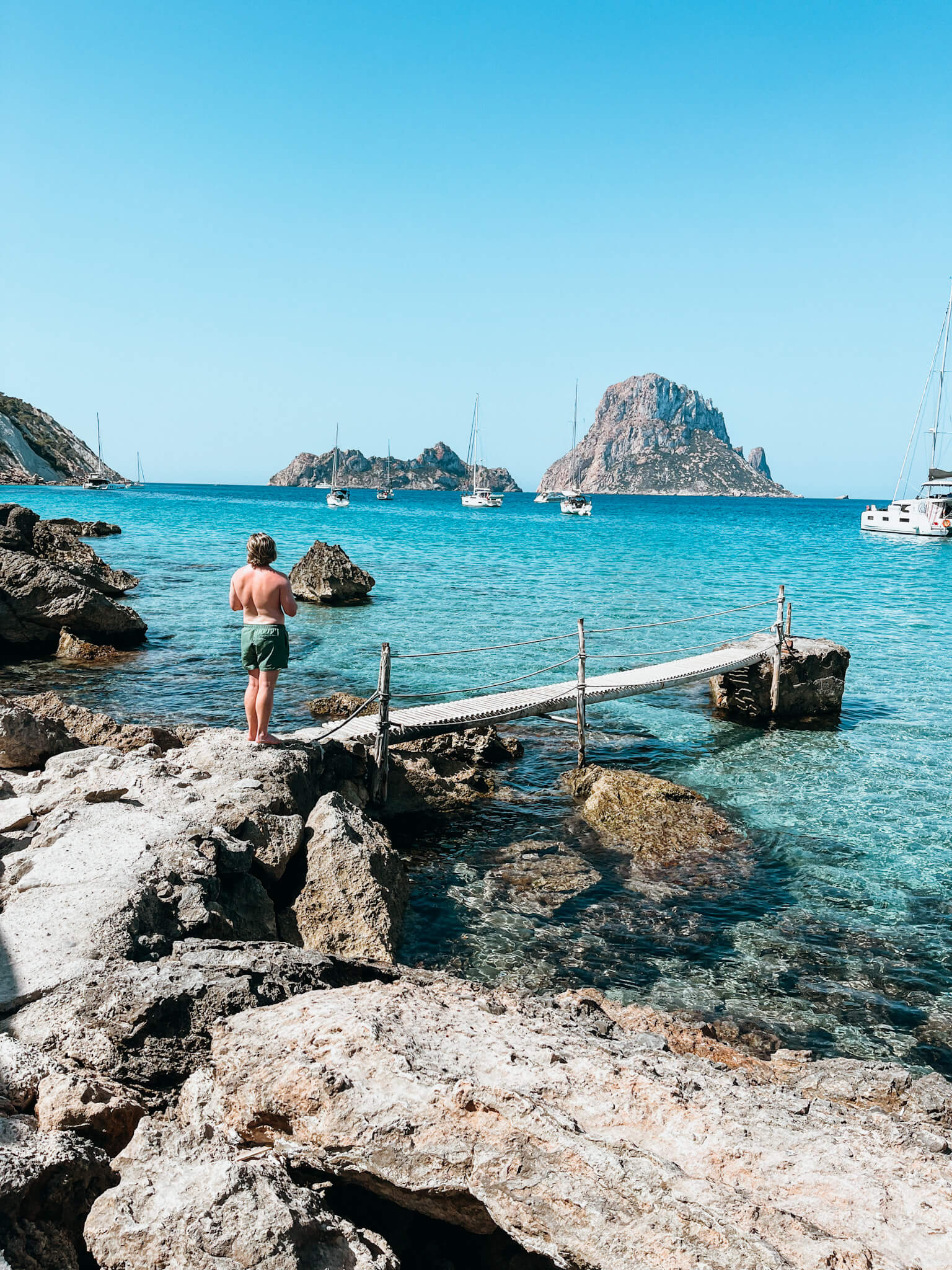 Man stands on small dock out into blue water with large rock in the background