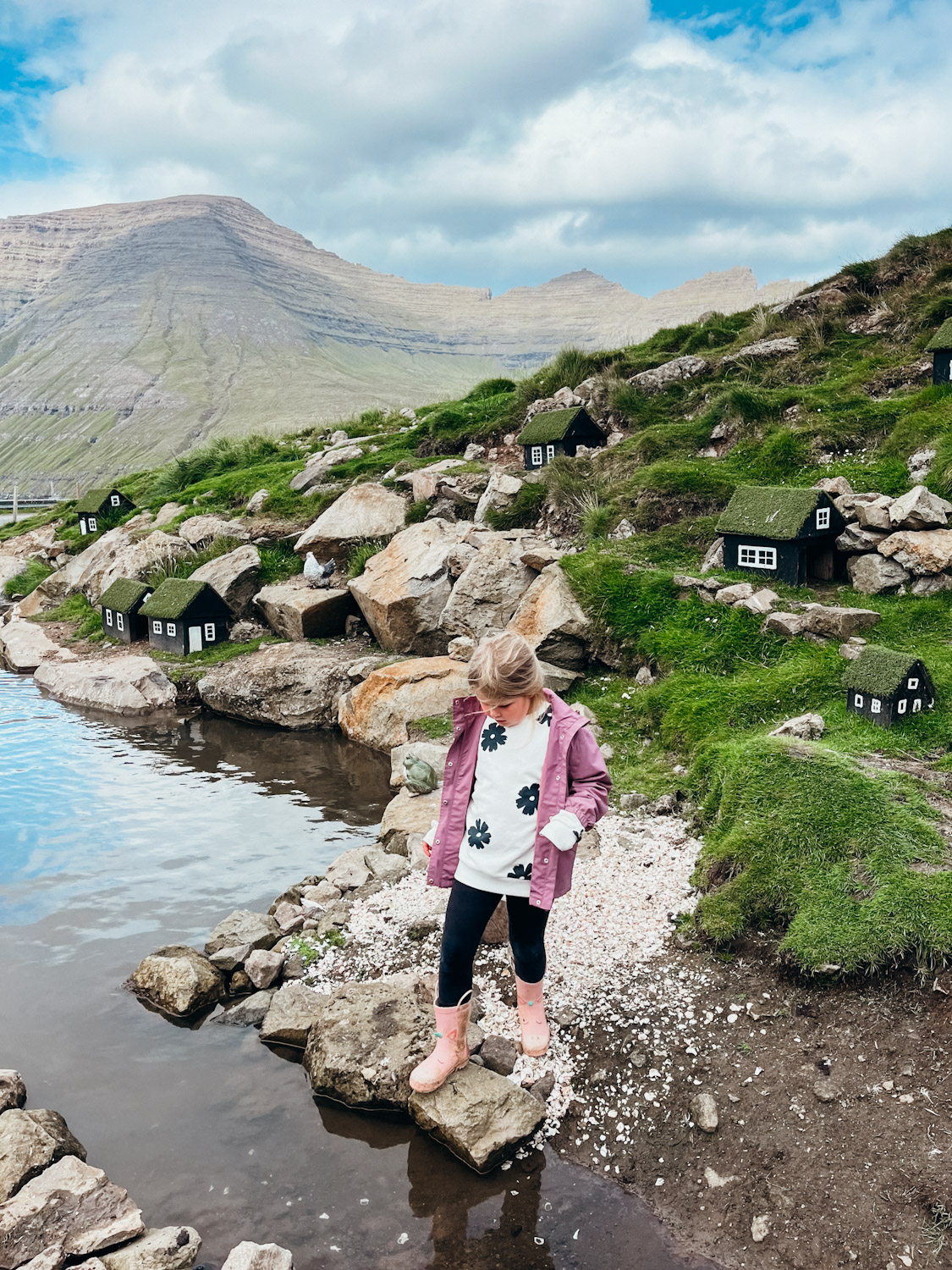 Child stands next to a pond with little birdhouses on the hill behind her