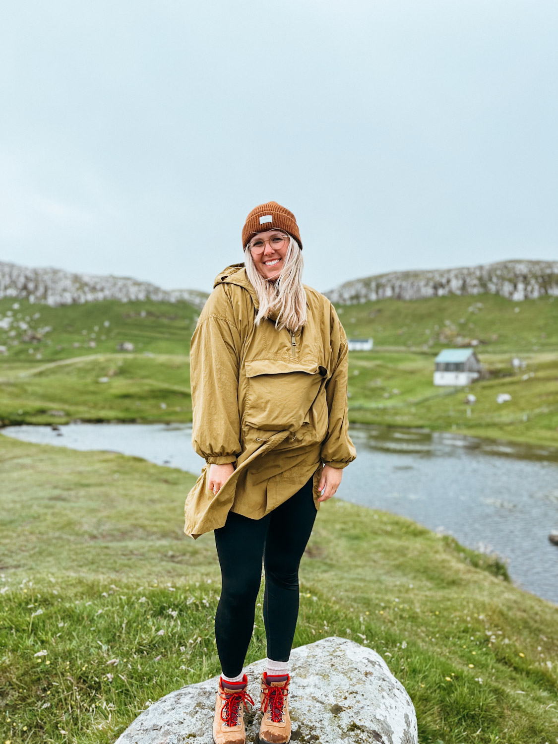 Woman stands on top of boulder with pond and sheep huts in the background