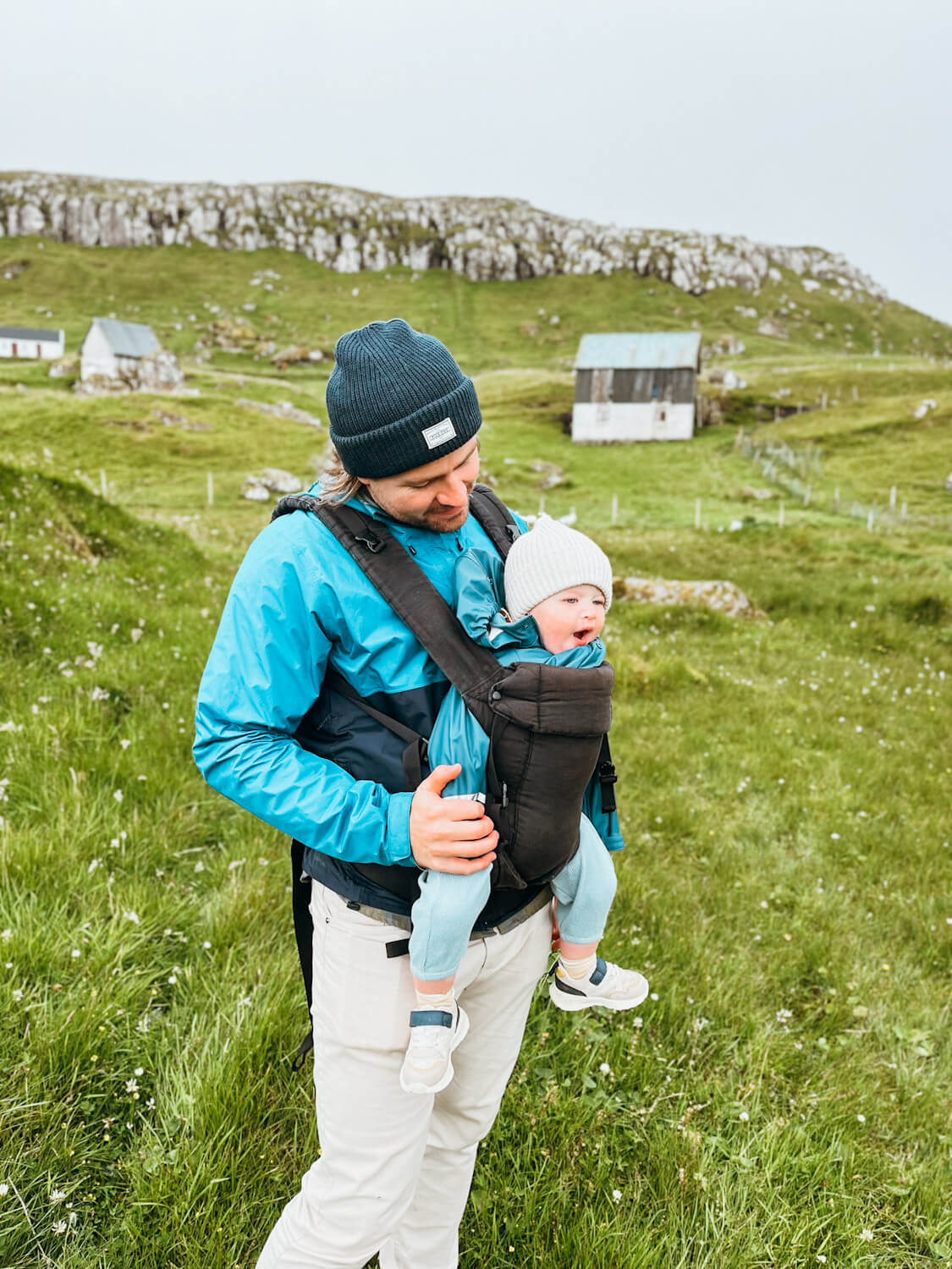 A man carries a baby in front of a green hill and old sheep huts