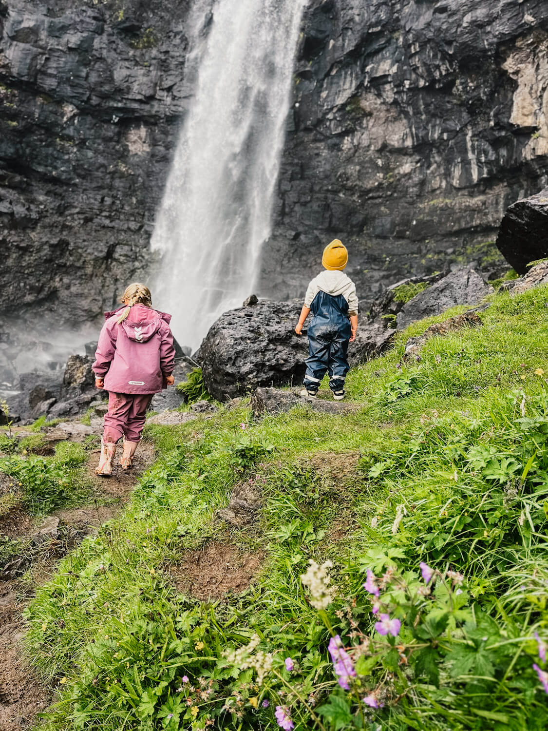 Two children stand facing a large waterfall in the Faroe Islands