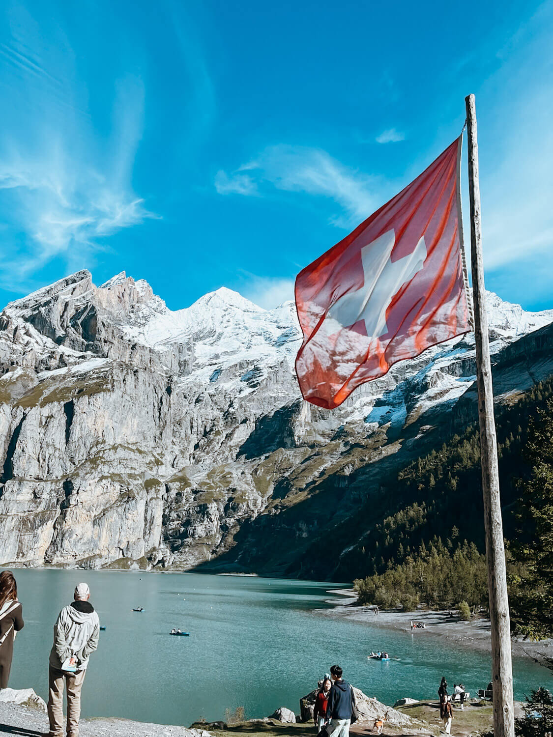 Red flag with white cross on wooden flag pool in front of a blue lake flanked by snow-capped mountains