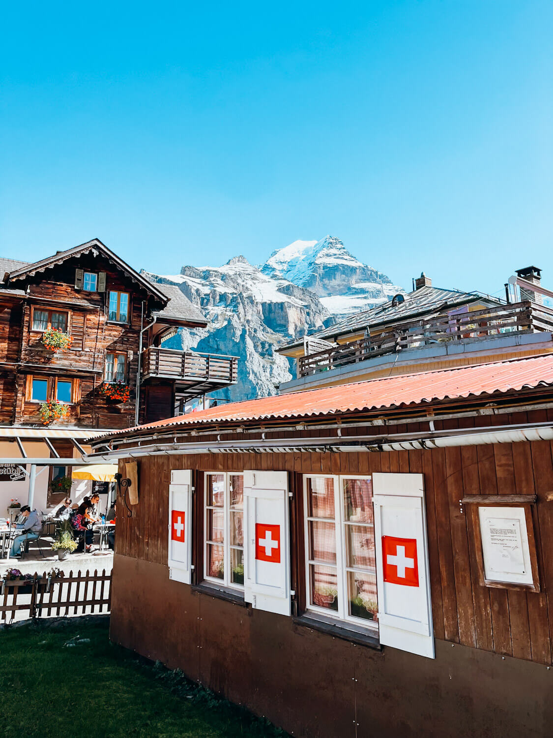 Wooden Swiss chalets with white shutters with red Swiss flags with large snow-covered peaks in the background in Mürren Switzerland