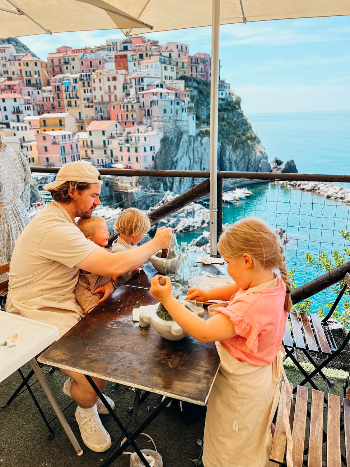 Father and three children make pesto at a wooden table with views of Manarola behind them at Nessun Dorma