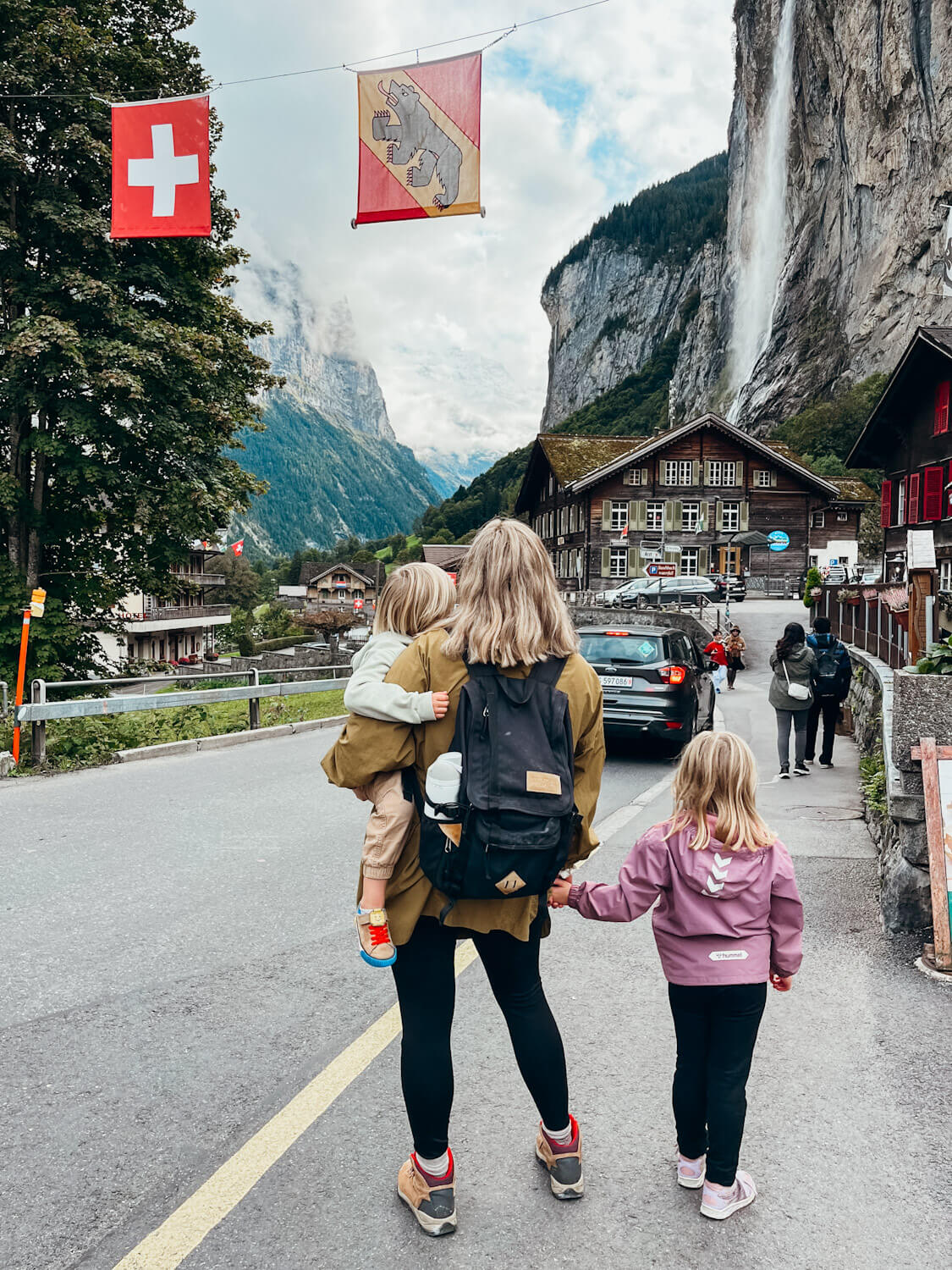 Woman and children stand on side of road with waterfall view and Swiss flags hanging overhead