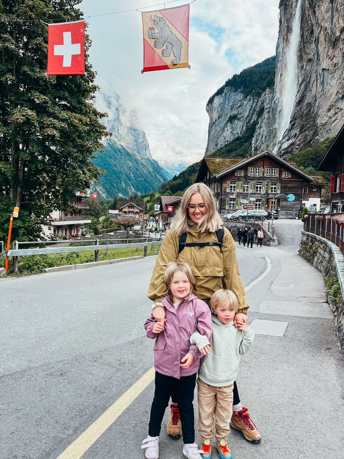 Woman and two children stand under Swiss flags with waterfall coming from cliffside in Lauterbrunnen with kids