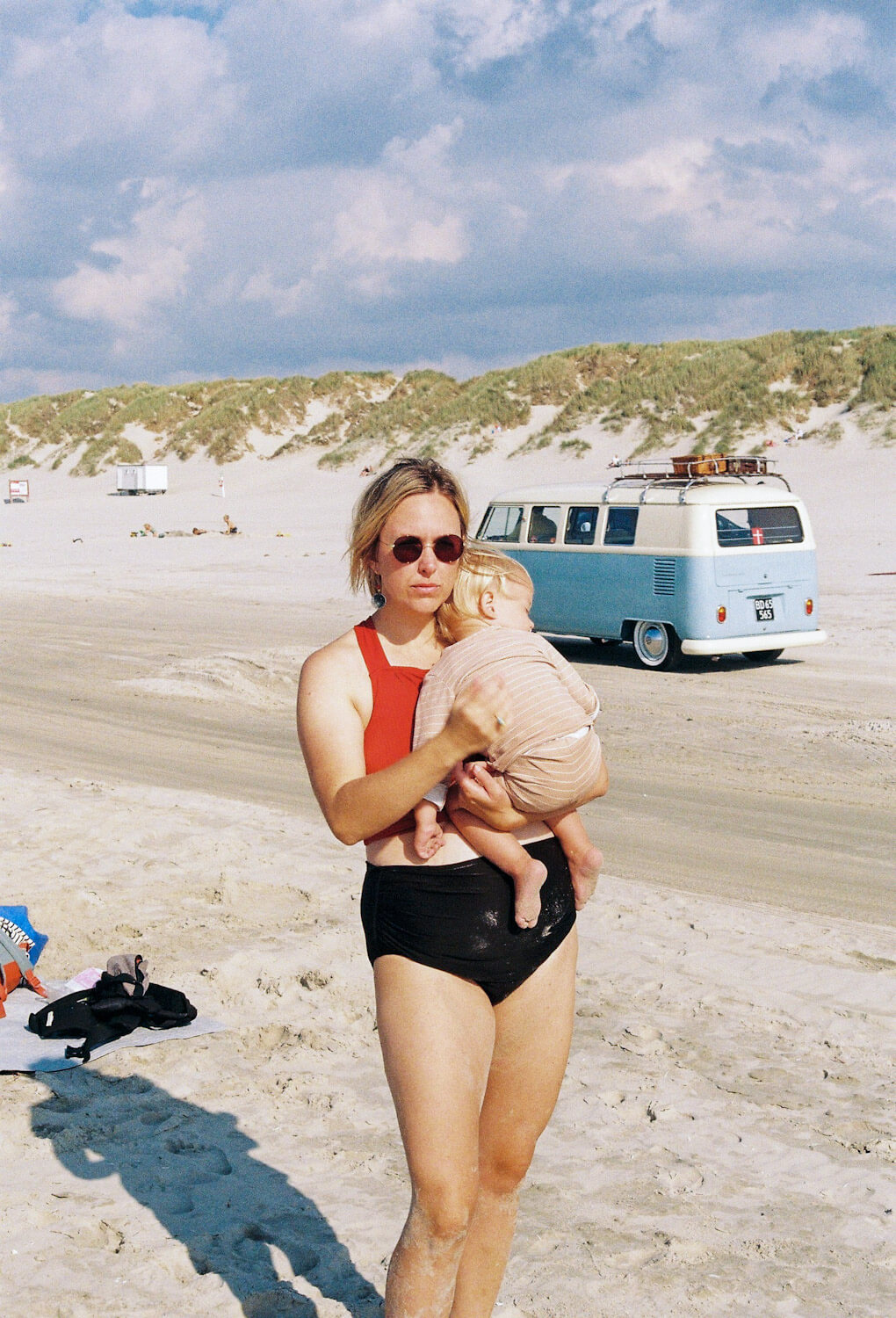 Woman holds child on long sandy beach while a blue and white VW van drives behind them at Vejers Strand Beach