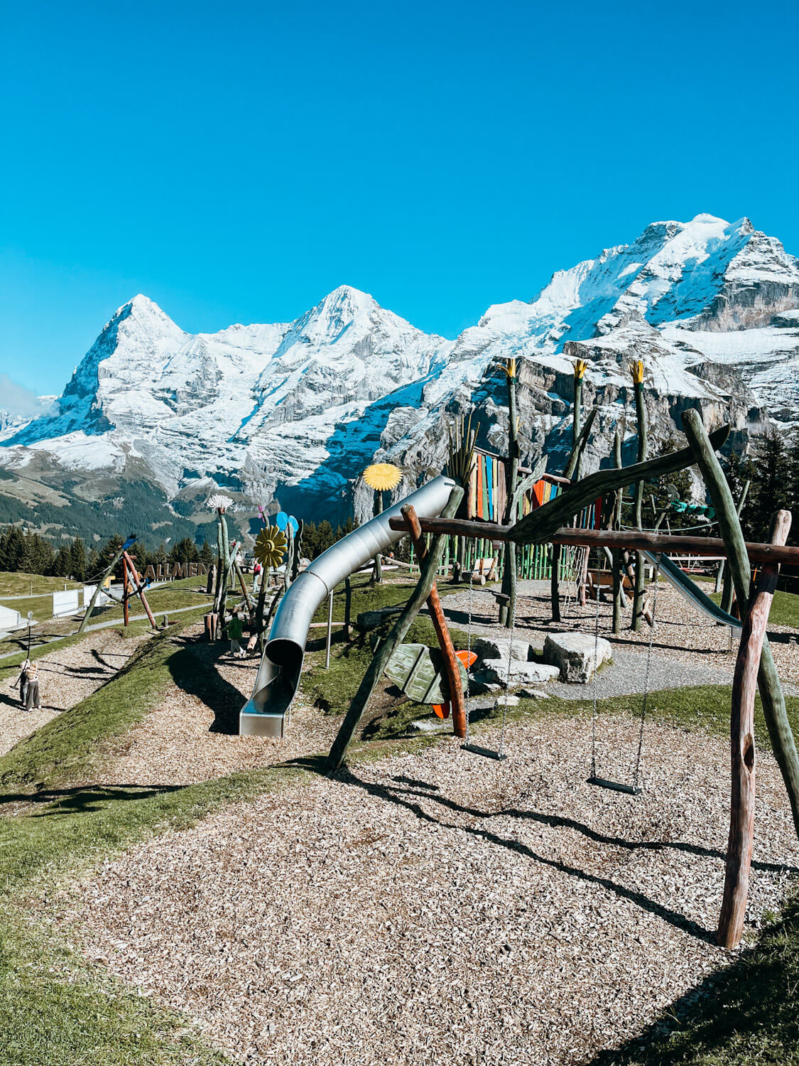 Wooden play structures at Allmendhubel Flower Park playground in Murren Switzerland with views of snow-capped mountain peaks