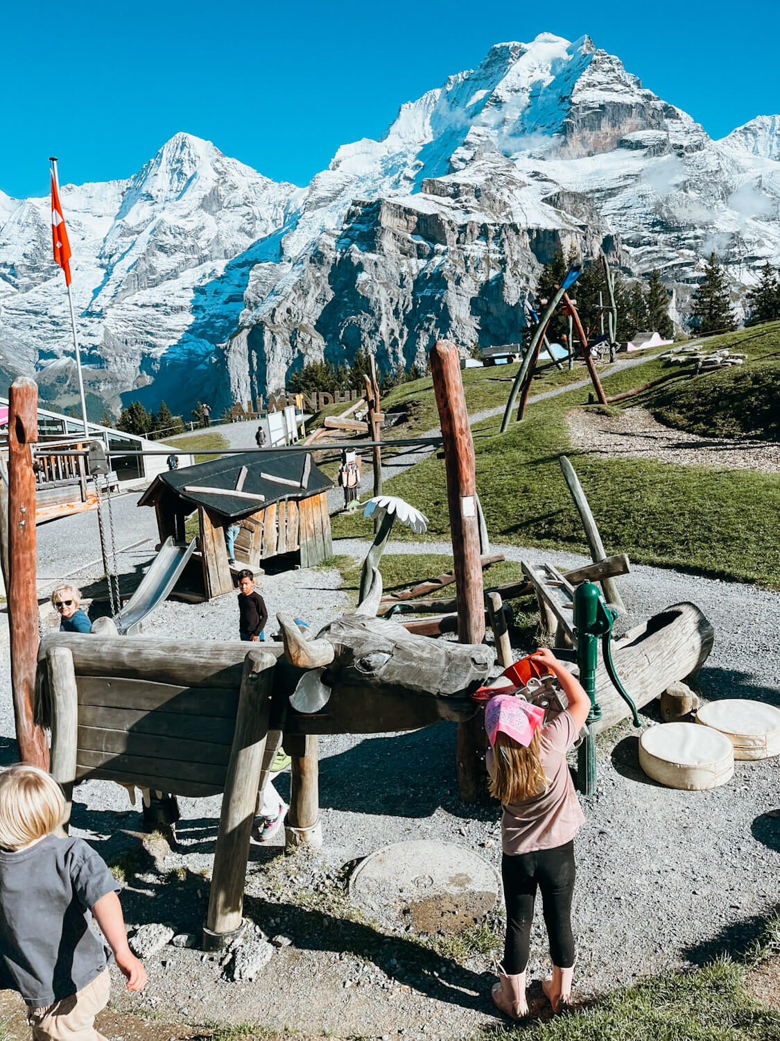 Wooden play structures at Allmendhubel Flower Park playground in Murren Switzerland with views of snow-capped mountain peaks