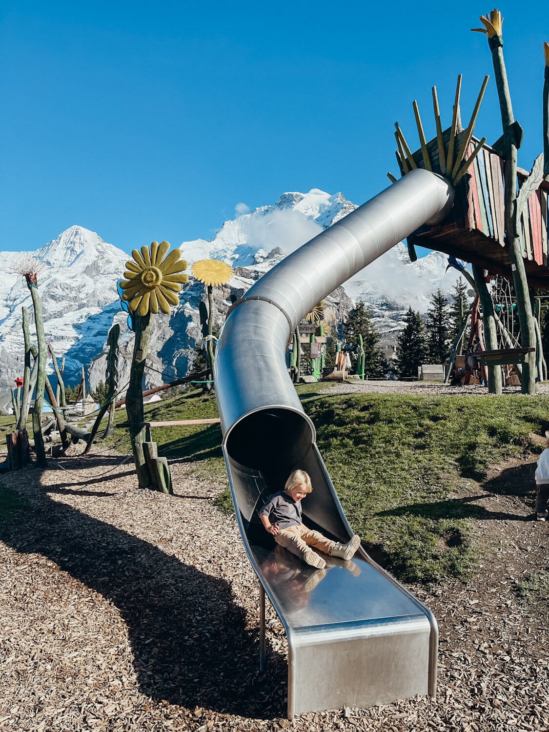 Child goes down a metal slide at Allmendhubel Flower Park playground in Murren Switzerland with views of snow-capped mountain peaks