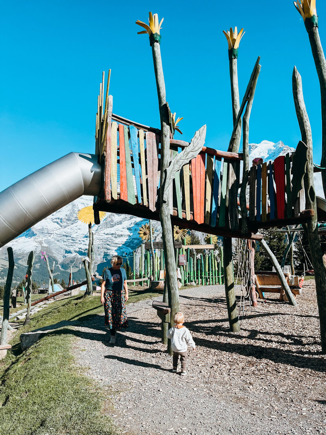 Wooden play structures at Allmendhubel Flower Park playground in Murren Switzerland with views of snow-capped mountain peaks
