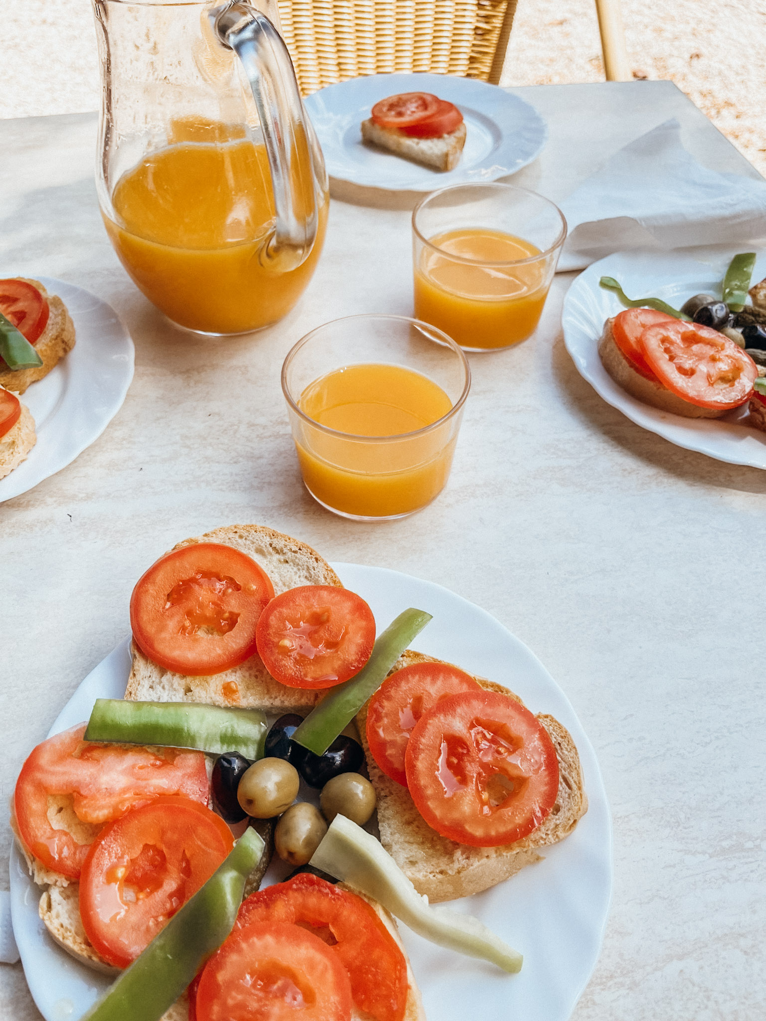 Plates of bread and tomatoes on a white table next to cups of orange juice