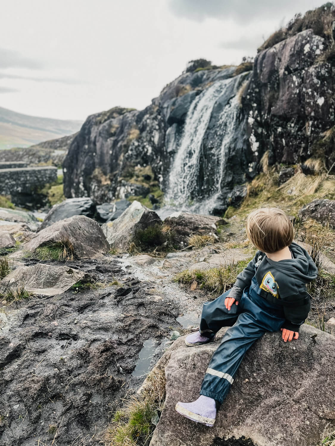 Child sits on rock in front of waterfall on Conor Pass in Dingle with children