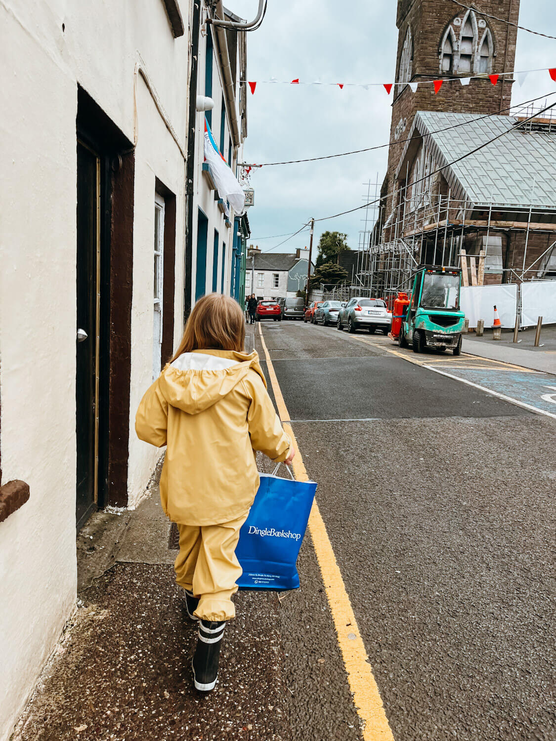 Girl in yellow rainsuit walkds down sidewalk holding blue 'Dingle Bookshop' shopping bag in Dingle Ireland with kids
