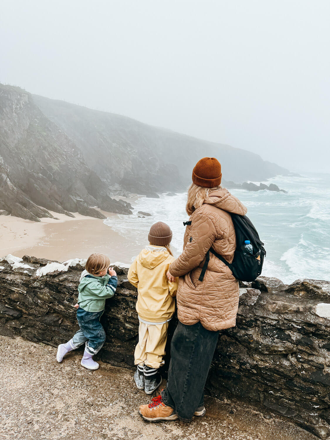 A woman and two children stand next to wall overlooking foggy, sandy beach in Dingle with kids