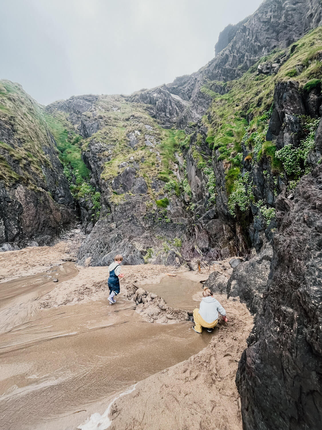 Two children play with sand next to a green, rocky cliff on a beach in Dingle Ireland with kids