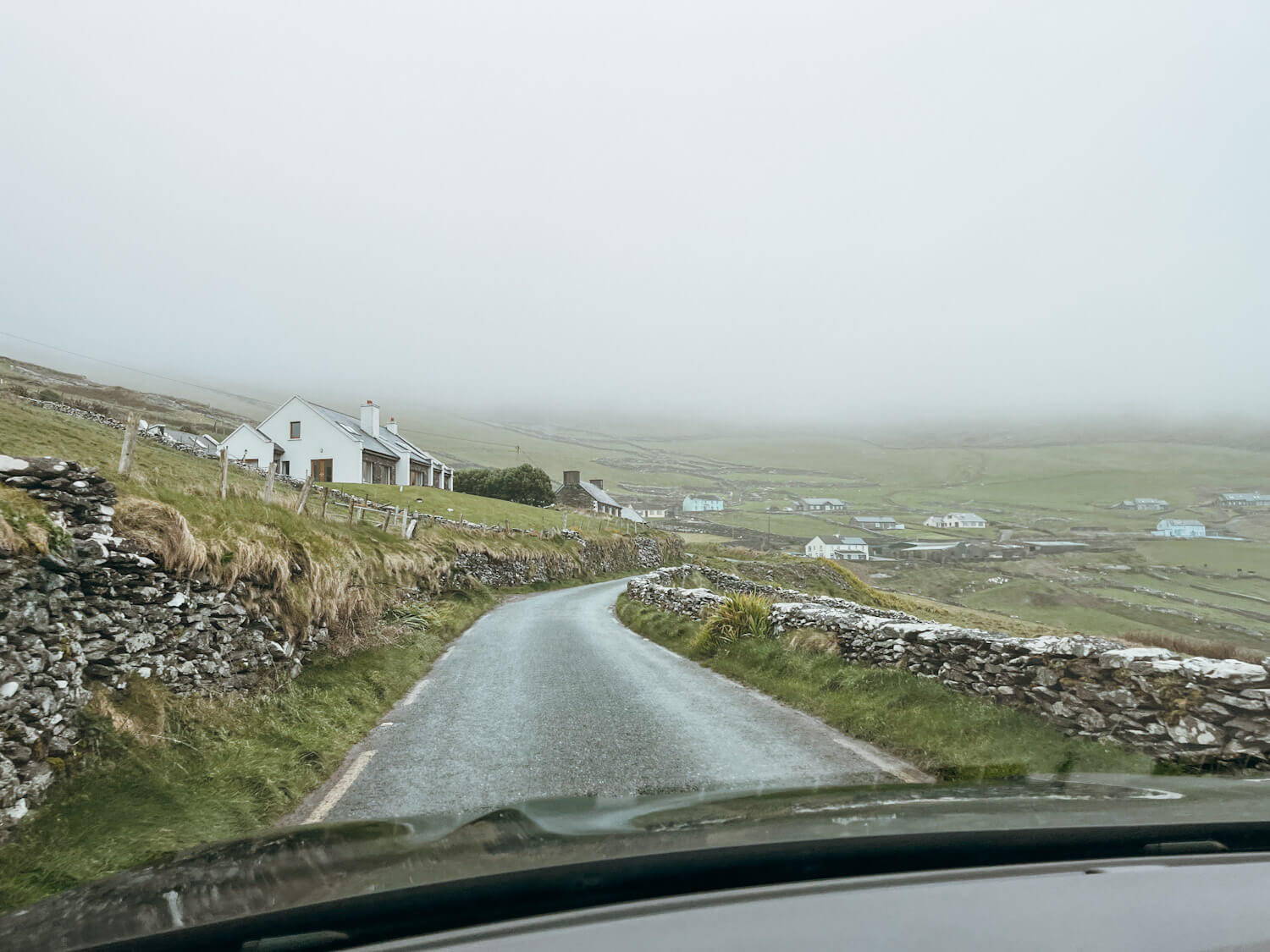 View from a windshield on a small road lined by green hill with white cottages