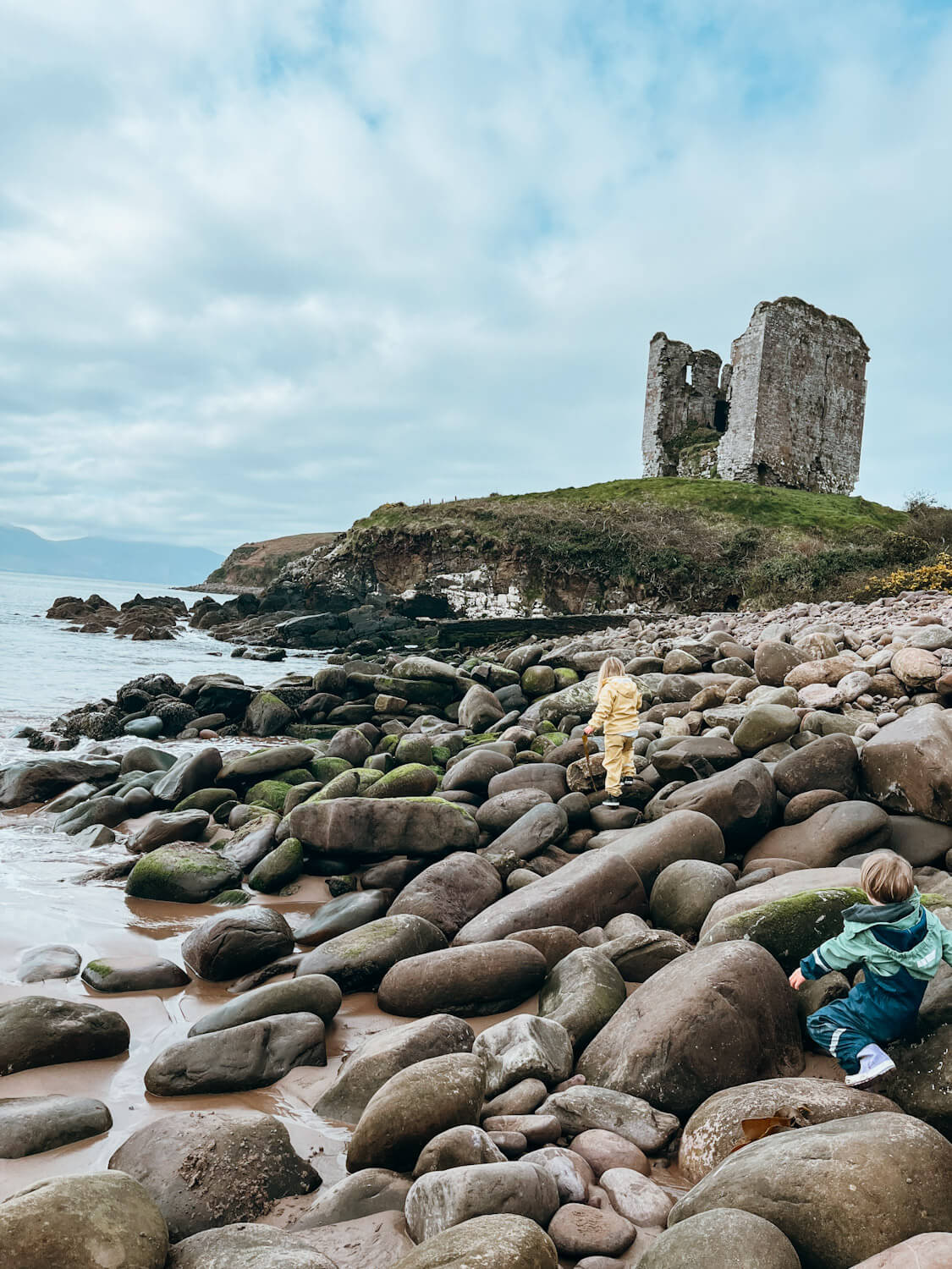 Two children scramble on boulders next to sea with castle ruins in the distance on a hill