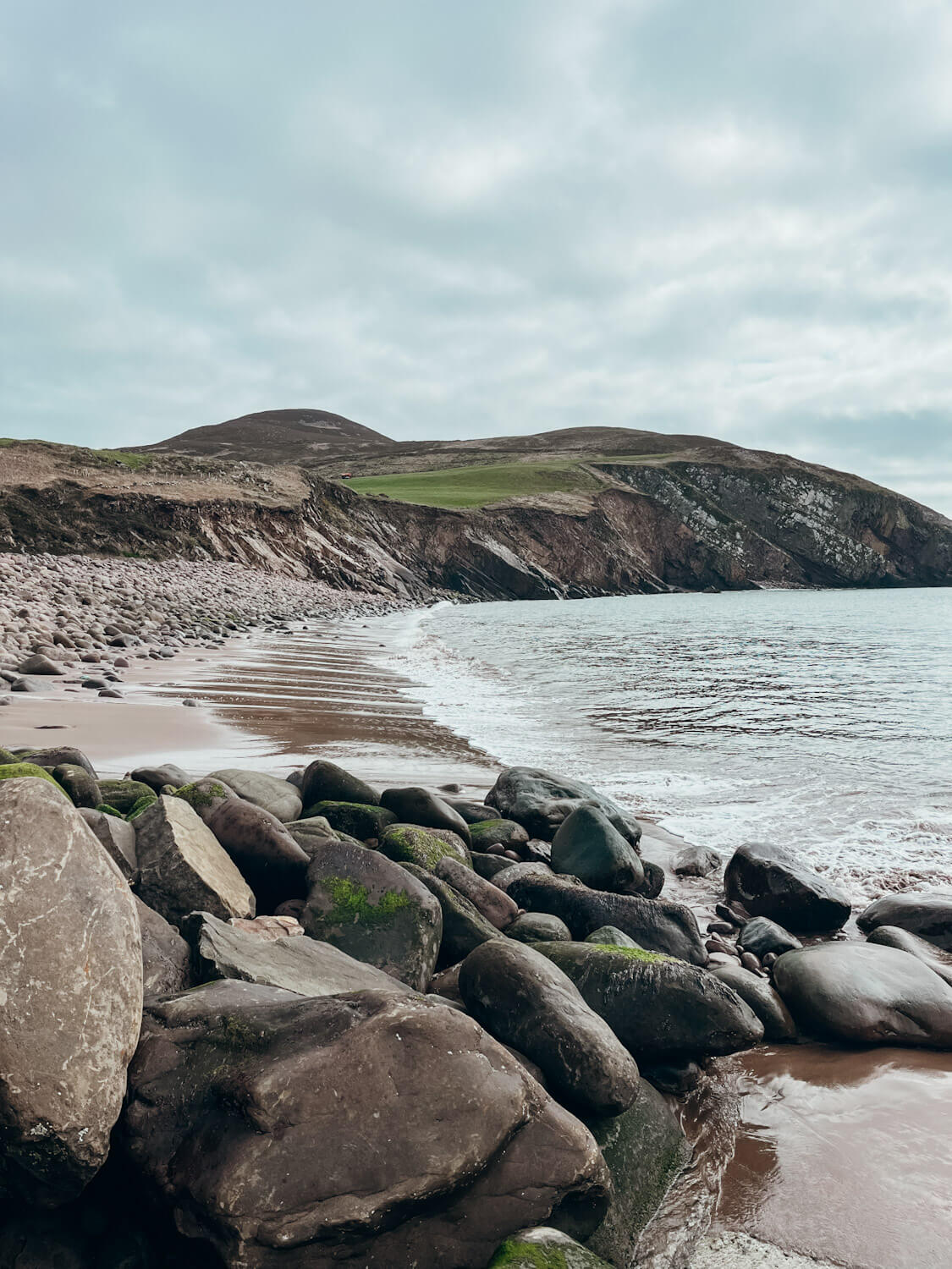 Sandy beach lined with large boulders with green cliffs in the distance