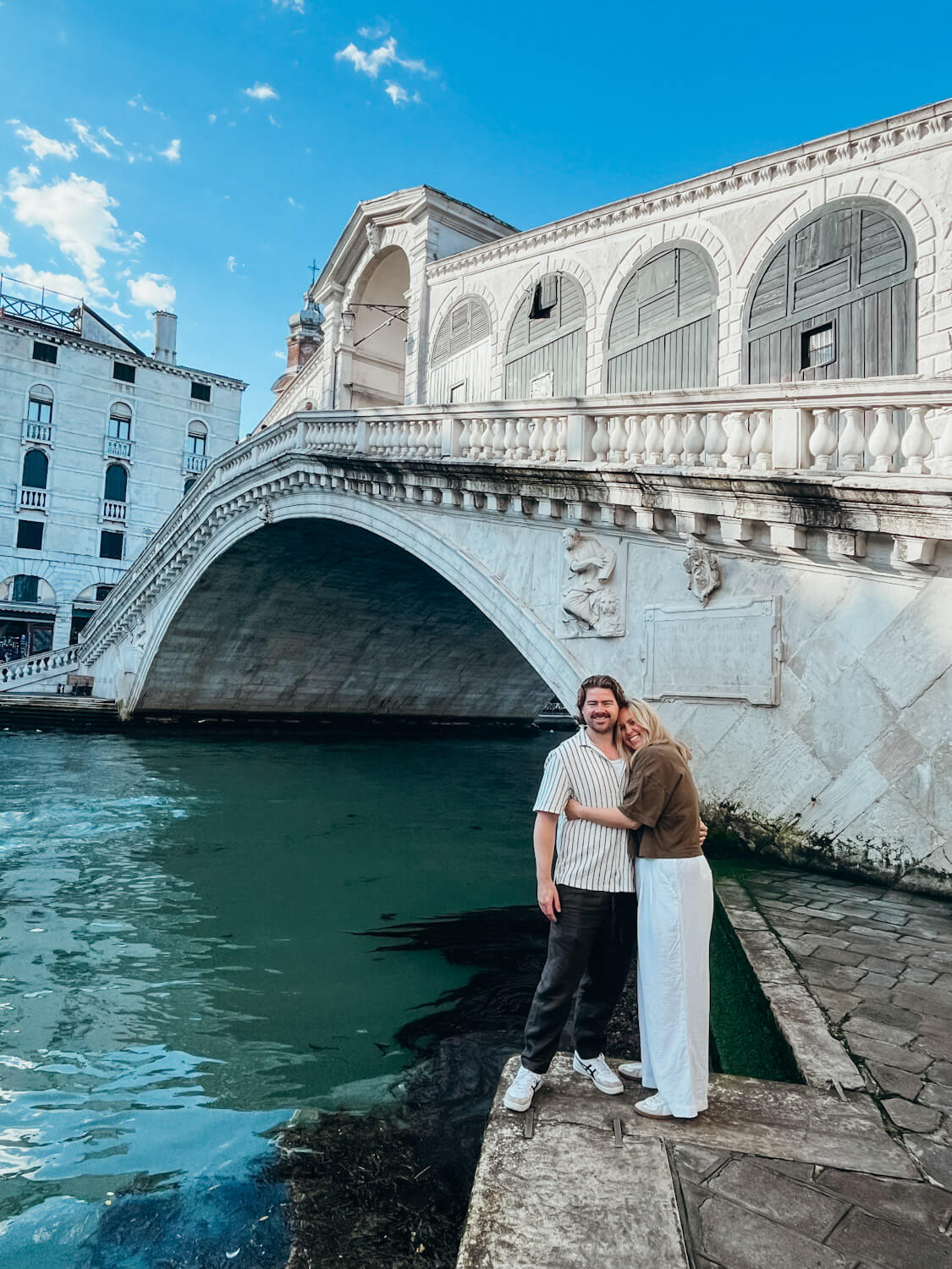 Woman and man hug in front of large white, stone bridge in May in Venice