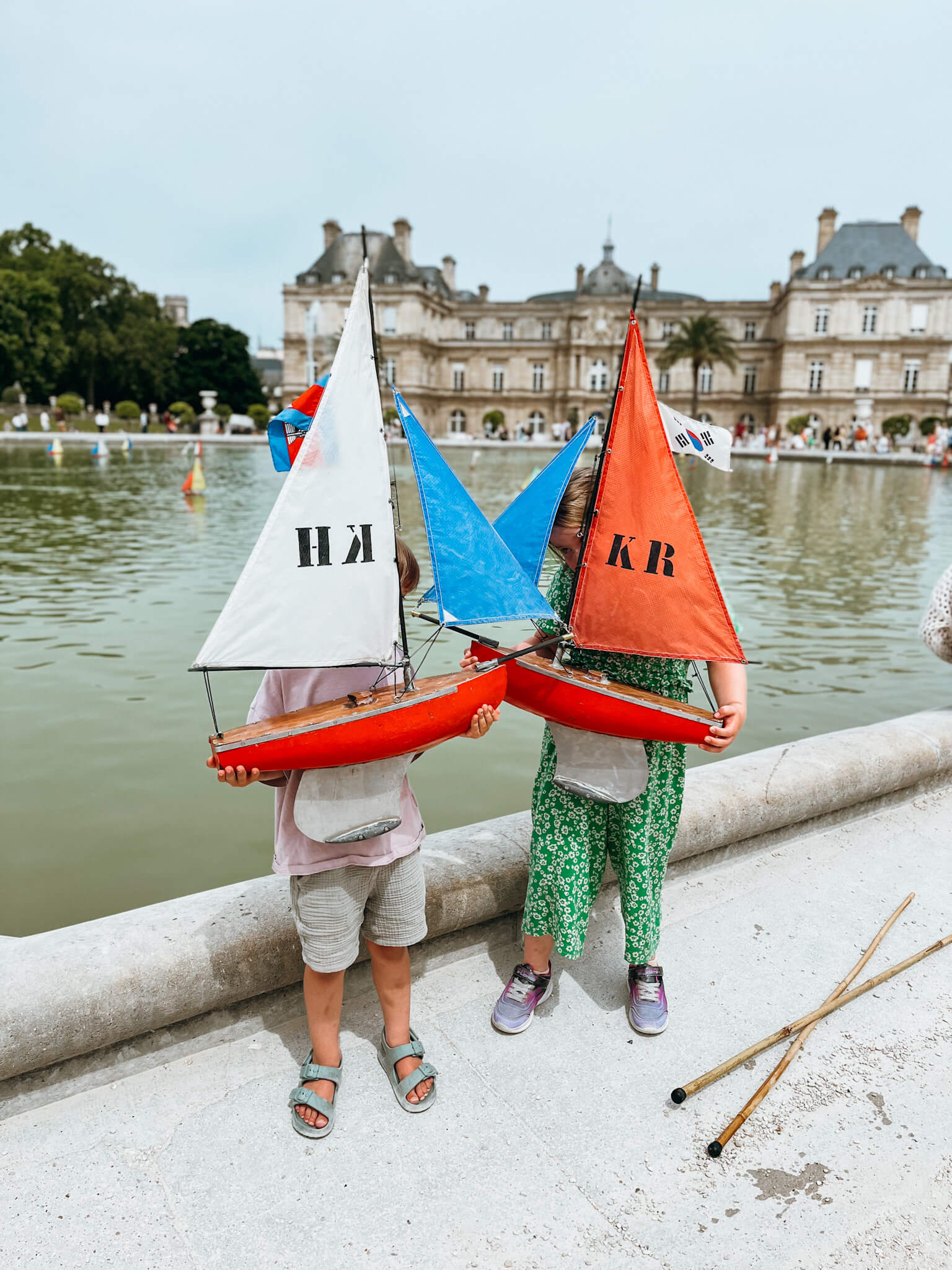 Two children hold vintage colorful toy sailboats in front of a fountain at Luxembourg Gardes in Paris in May