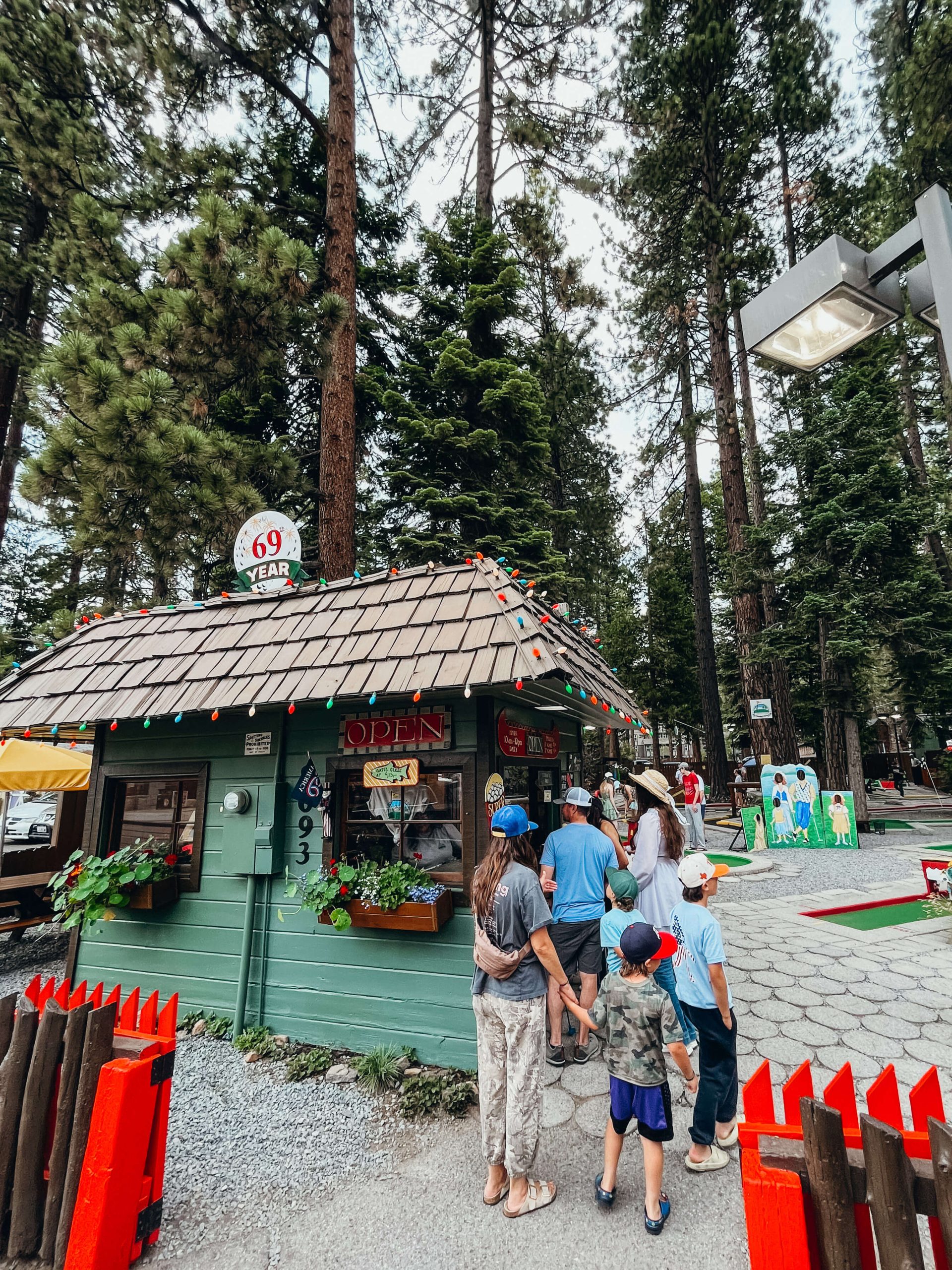 People stand in a line next to a small, green shack next to a mini golf course to pay for entrance