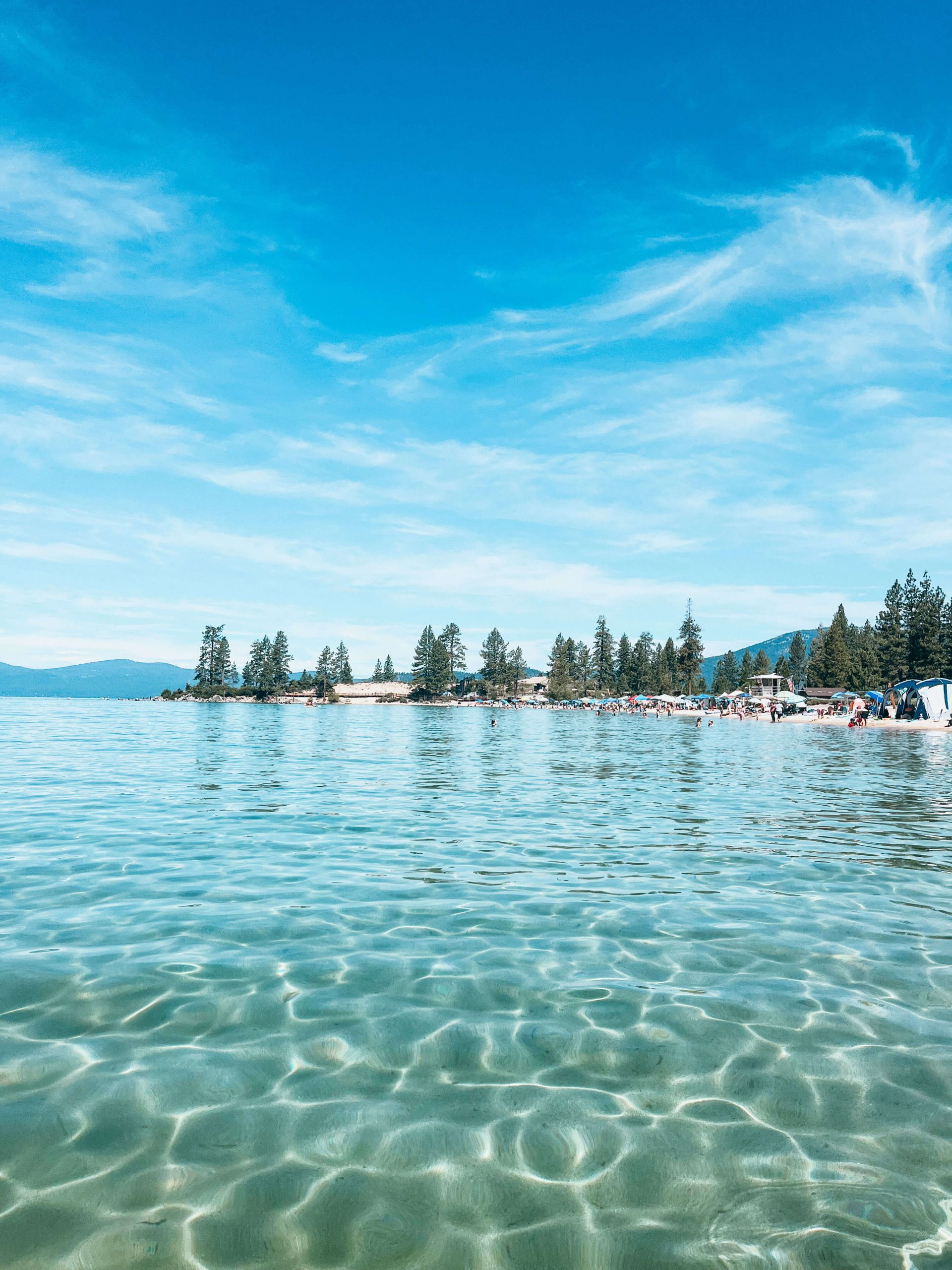 Blue and green water next to sandy beach lined with pine trees and umbrellas at Sand Harbor State Park