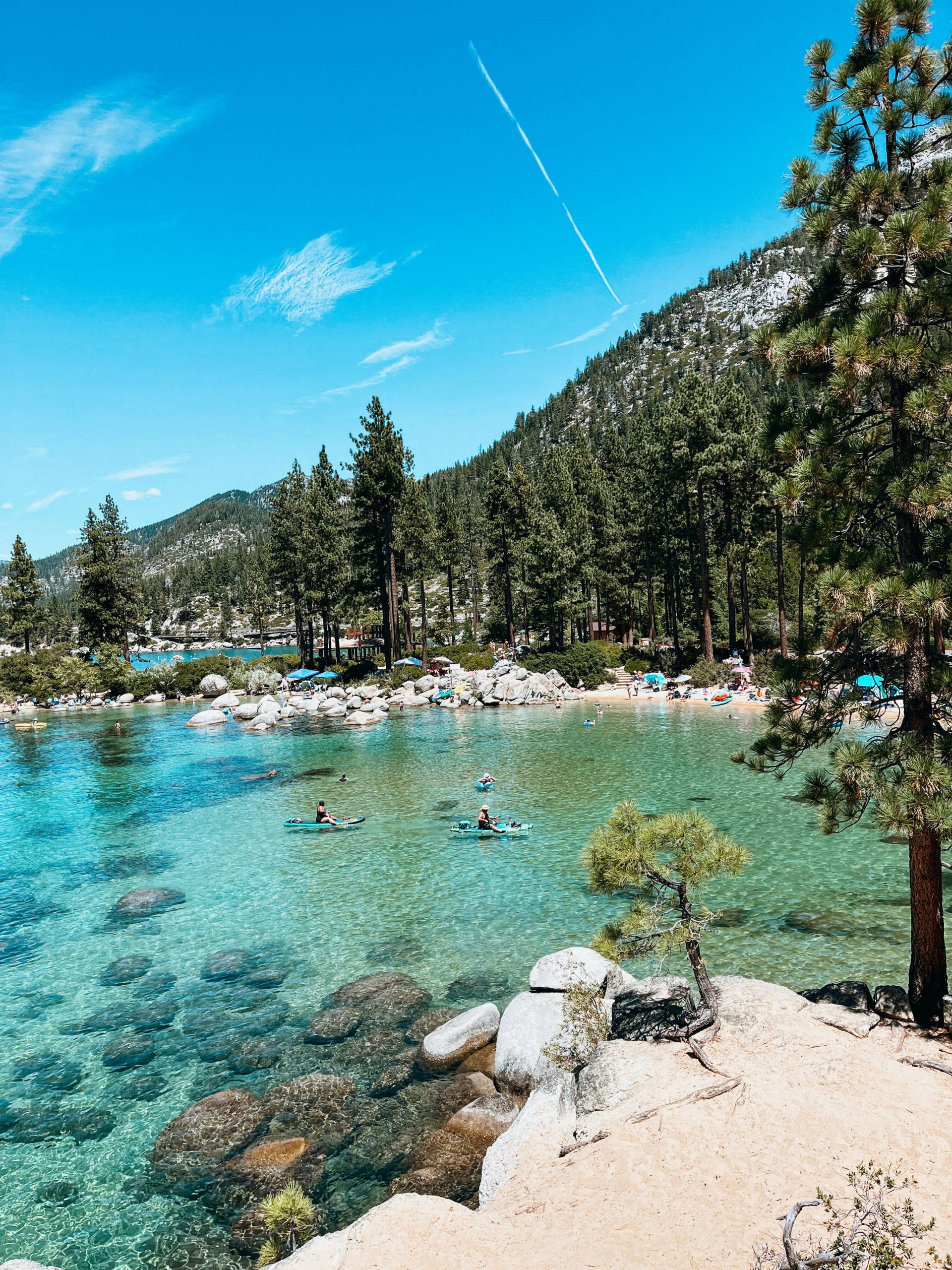 Sandy cove lined with boulders and pine trees with crystal clear blue and green water with kayakers at Sand Harbor Beach