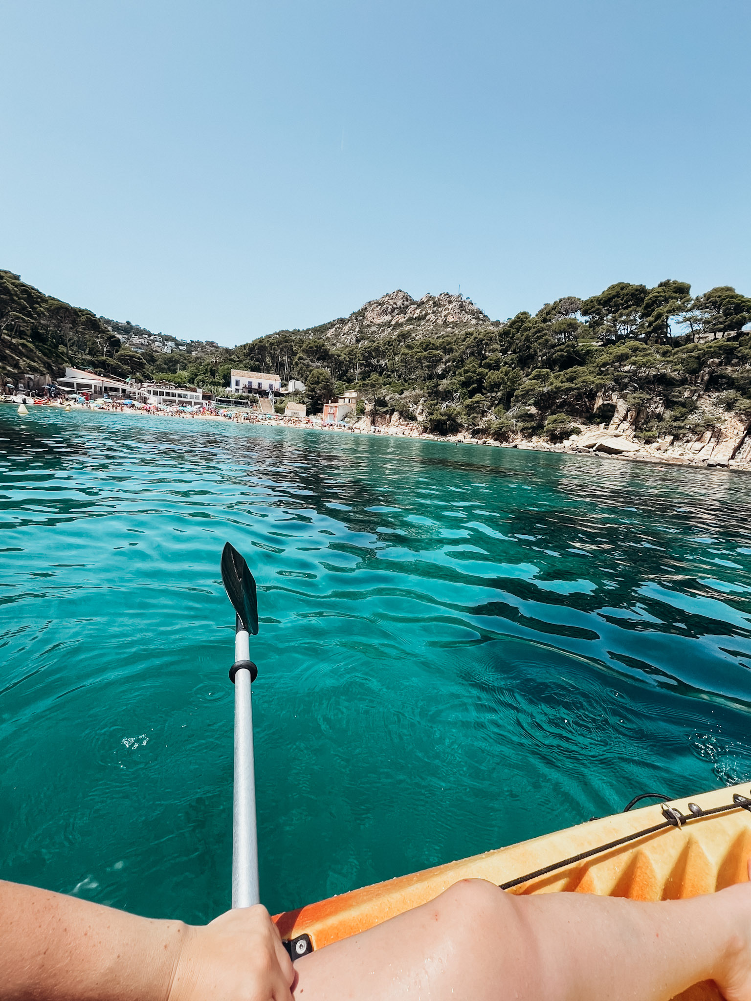 Image of edge of kayak and paddle over turquoise water with beach in the distance at Aiguablava