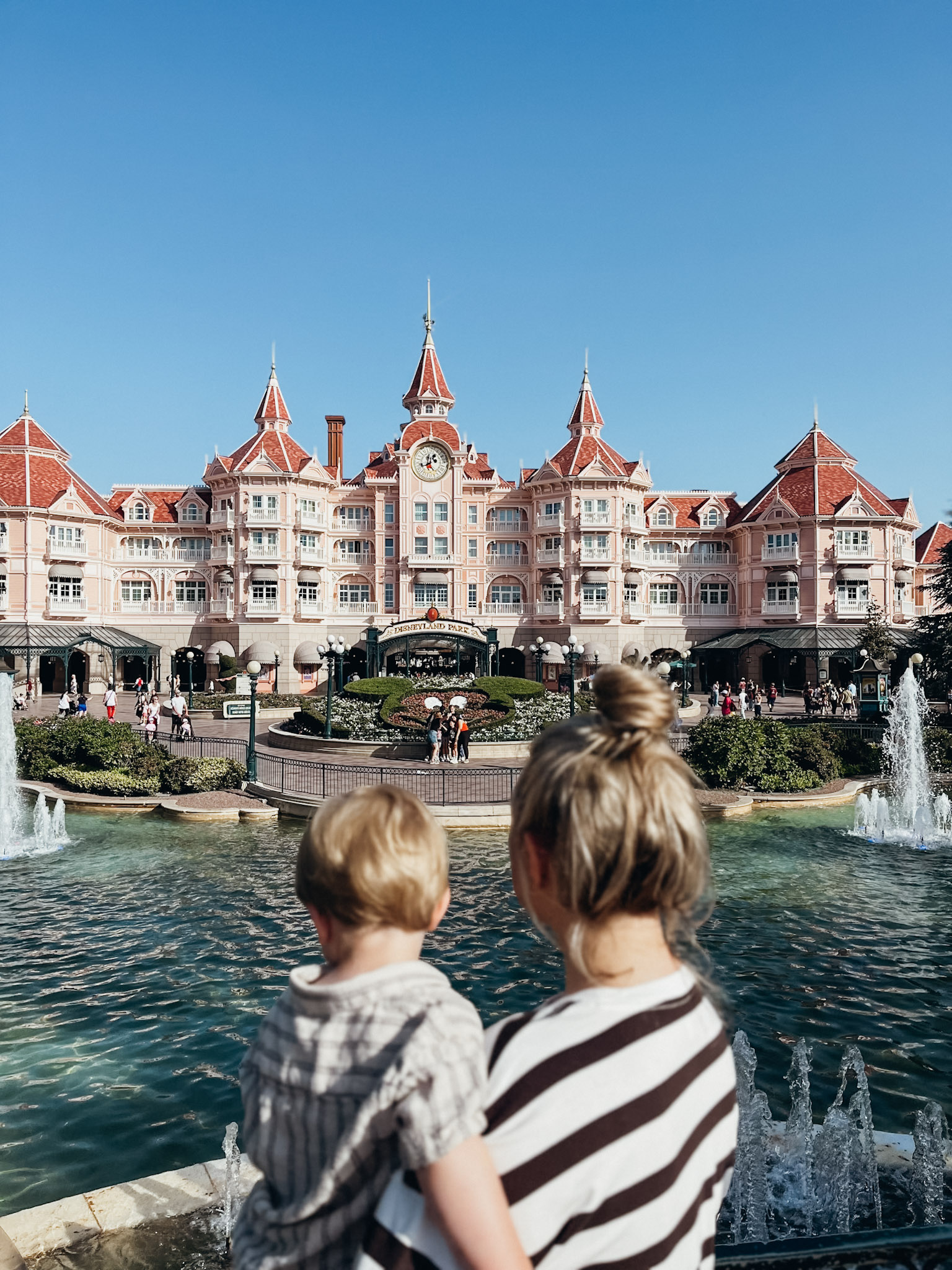 Woman and child stand in front of fountain and large pink building at Disneyland Paris in May