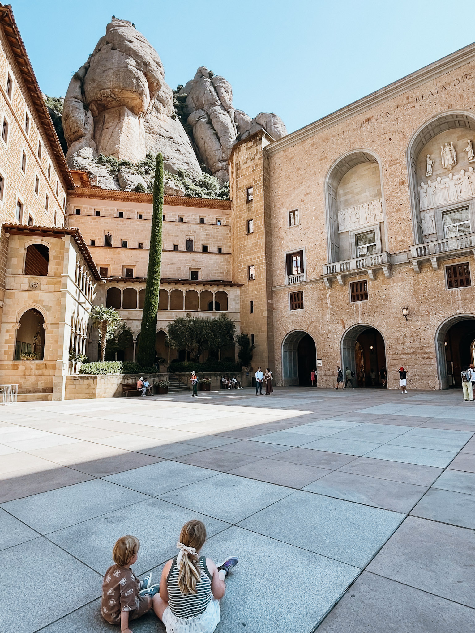 Two young children sit on stone in front of monastery built in to the mountain at Montserrat near Barcelona in June
